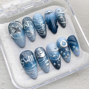 May include: A set of ten blue and silver press-on nails. Each nail features a unique design, including text, stars, and wave patterns. The nails are displayed in a clear plastic container.