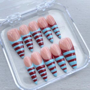 Op de afbeelding: Een set van tien kunstnagels in een doorzichtige plastic doos. De nagels zijn amandelvormig met een huidkleurige basis en een ontwerp met afwisselende lichtblauwe en kastanjebruine horizontale strepen. De punten zijn puntig.