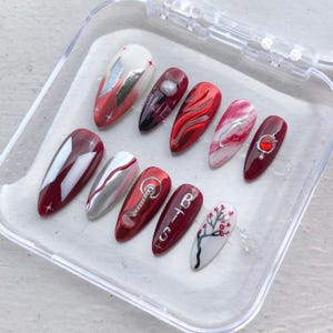 May include: A set of ten artificial nails in a clear plastic case. The nails feature various designs, including silver foil accents, abstract patterns, and a cherry blossom design. Colors include red, white, and silver.