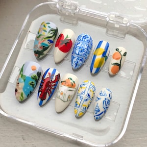 May include: A set of ten almond-shaped false nails, each hand-painted with unique designs. The designs include landscapes, a lobster, fruit, and geometric patterns in various colors, set in a clear plastic case.