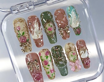 May include: A set of ten press-on nails in a clear plastic case, featuring various designs including pink roses, white swans, gold bows, and pearl accents. The nails are painted in shades of cream, dusty rose, sage green, and mint green with 3D details.