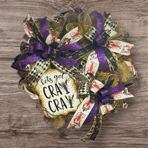 May include: A decorative wreath with purple, gold, and black ribbons. The wreath features a sign that reads "lets get CRA CRA" with crawfish illustrations. The wreath is made of mesh and has multiple bows.