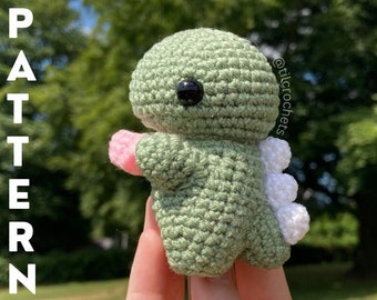 May include: A green crocheted dinosaur toy with white spikes on its back and pink arms. The toy is being held by a hand.
