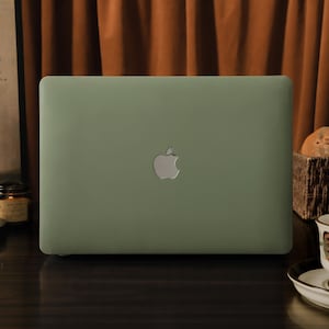 Peut inclure: Un ordinateur portable vert sauge avec un logo Apple argenté. Il est posé sur une surface en bois sombre. Une tasse à thé et une soucoupe à motifs floraux sont à droite. Une peinture et un panier de pain sont en arrière-plan.