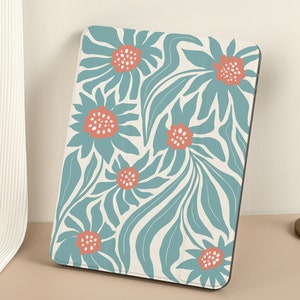 Puede incluir: Una funda para iPad con estampado floral en azul verdoso y blanco con un diseño de flores estilizadas. Las flores tienen un centro rosa coral.