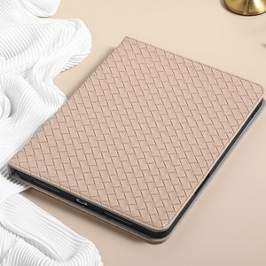 May include: A light beige woven leather iPad case with a black trim. The case is lying on a white fabric surface.