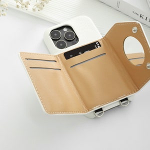 May include: White leather phone case with a built-in wallet and a small mirror. The case has three card slots and a strap for carrying.