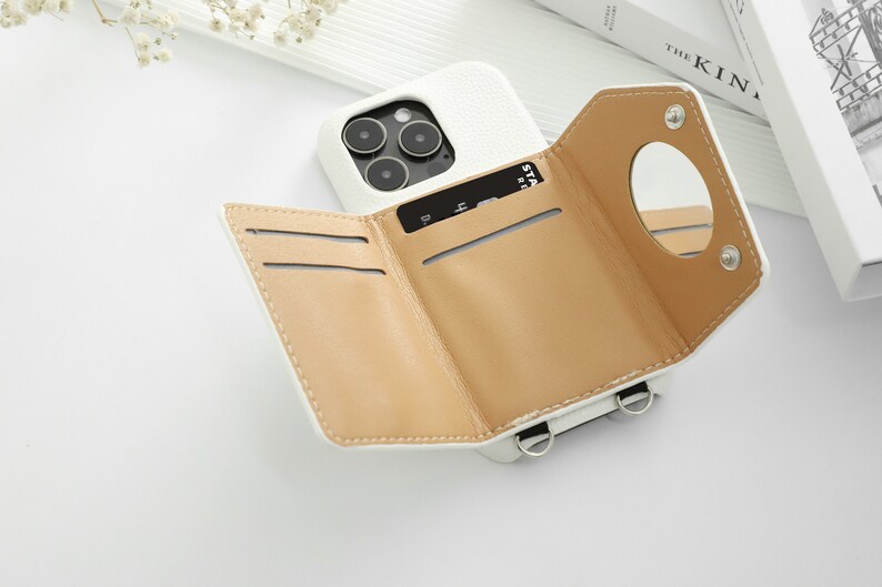 May include: White leather phone case with a built-in wallet and a small mirror. The case has three card slots and a strap for carrying.