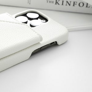 May include: A white leather phone case with a built-in wallet flap. The case is designed for a phone with a triple camera system.
