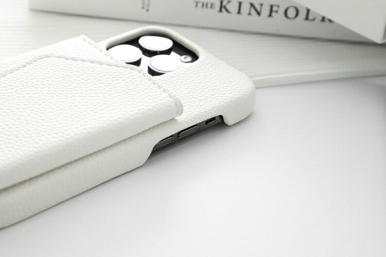 May include: A white leather phone case with a built-in wallet flap. The case is designed for a phone with a triple camera system.