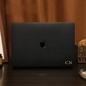 May include: A black laptop with the Apple logo and gold initials "C.N" is displayed on a dark wooden surface. The background includes a framed abstract art piece, candles, a basket of bread, and a teacup.