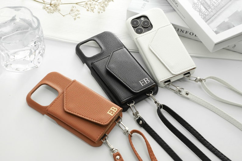 May include: Three leather phone cases with a pocket and a strap. The cases are brown, black, and white. Each case has the initials "EB" embossed on the front.