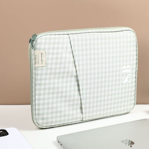 Puede incluir: Una funda para portátil de cuadros vichy verde y blanco con cierre de cremallera. La funda tiene un bolsillo en la parte delantera y las iniciales "SB" bordadas en el lado derecho. El texto "Early Work" está impreso en una pequeña etiqueta adjunta a la cremallera.