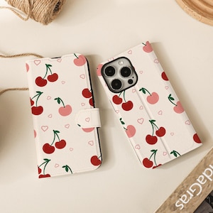 Peut inclure: Coque de téléphone blanche ornée d'un motif de cerises rouges et roses et de petits cœurs roses. L'étui est ouvert, révélant l'appareil photo et l'écran du téléphone. Un étui fermé assorti est également présent.