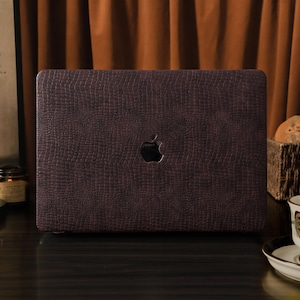 May include: A dark burgundy laptop case with a textured, reptile-like pattern. The case features a cutout for the Apple logo. The background includes a painting, a candle, and a teacup on a saucer.