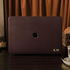 May include: A dark burgundy laptop with the Apple logo and gold initials "K.M." is displayed. The laptop is on a dark surface, with a teacup and saucer, a basket of bread, and a painting in the background. The background is a brown curtain.
