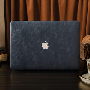 May include: A navy blue laptop case with a textured finish and a white Apple logo. The case is centered on a dark wooden surface. A cup of tea and a basket of bread are in the background.