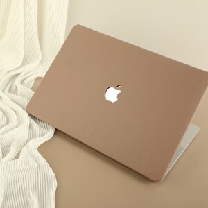 May include: A light brown laptop case with a textured surface and a white Apple logo. The case is closed and sitting on a light brown surface.