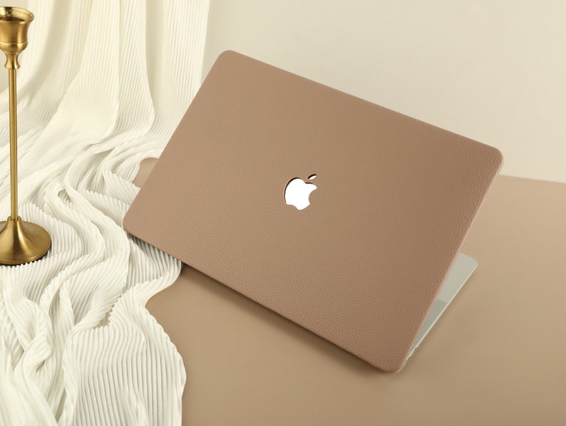 May include: A light brown laptop case with a textured surface and a white Apple logo. The case is closed and sitting on a light brown surface.
