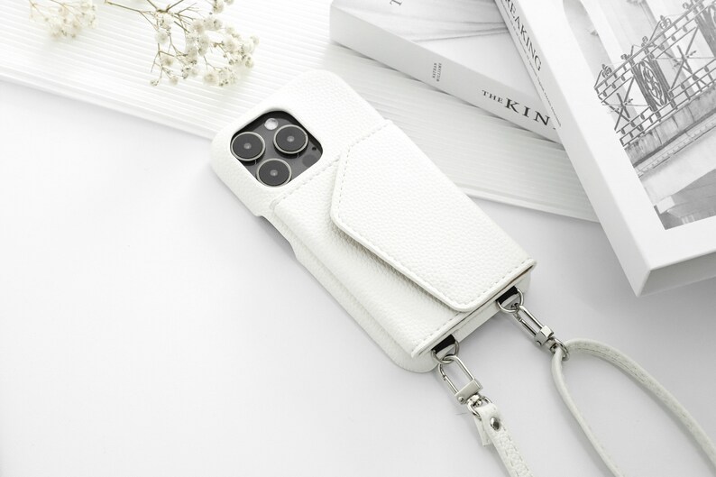 May include: White leather phone case with a flap and a white strap. The case has a camera lens cutout and a silver metal clasp.