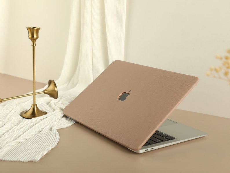 May include: A light brown leather laptop case with the Apple logo on the front. The case is closed and the laptop is visible underneath.