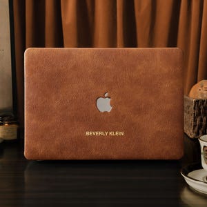 May include: A brown leather laptop case featuring the Apple logo and the name "BEVERLY KLEIN" in gold. The case is on a dark surface, with a cup of tea and a basket of bread in the background.