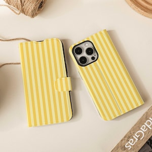 Peut inclure: Deux étuis de téléphone à rayures jaunes et blanches. Un étui est ouvert, révélant l'appareil photo du téléphone. L'autre étui est fermé avec une fermeture magnétique. Les étuis sont sur une surface blanche avec une pelote de ficelle et un plateau en bois.