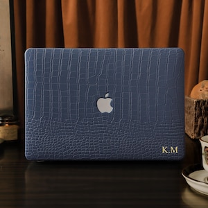 May include: A navy blue laptop case with a textured, crocodile-like pattern. The Apple logo is visible, and gold initials "K.M" are at the bottom right. The case is on a dark surface, with a cup of tea and bread in the background.