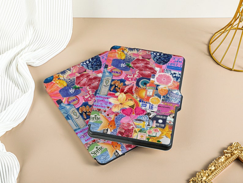May include: Two black e-reader cases with a colorful collage design. The design features pink lips, a blue eye, a pineapple, a papaya, a lime, a grapefruit, a gin bottle, a Sprite can, a fish, and the words 'Rima', 'Love Forever', 'Lanai', 'Don't Stop', 'Vogue', 'Gardine', and 'Cava'. 