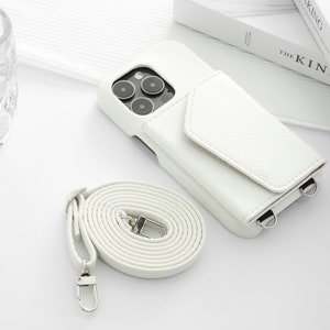 May include: White leather phone case with a flap and a detachable strap. The case has a camera cutout and two metal rings for attaching the strap.