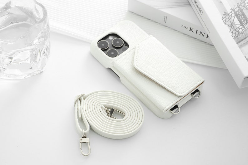 May include: White leather phone case with a flap and a detachable strap. The case has a camera cutout and two metal rings for attaching the strap.