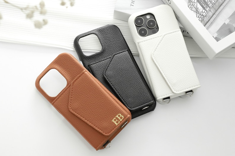May include: Three phone cases with a pocket on the back. The cases are brown, black, and white. The brown case has the initials "EB" embossed on the pocket. All cases have a strap.