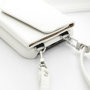 May include: White leather phone case with a flap closure and a detachable strap. The case has a silver metal clasp and a silver metal ring.