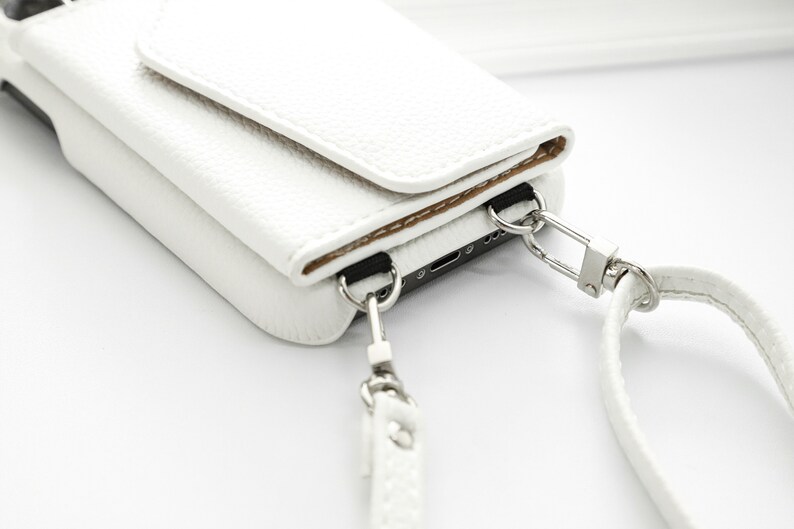 May include: White leather phone case with a flap closure and a detachable strap. The case has a silver metal clasp and a silver metal ring.