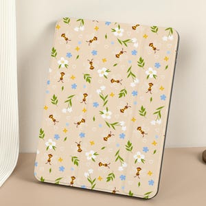 Pode incluir: Uma capa para iPad bege com um padrão de flores brancas, folhas amarelas e abelhas marrons.