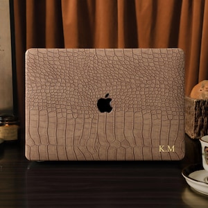 May include: A tan laptop case with a crocodile skin texture. The case has a black Apple logo and gold initials "K.M." on the lower right corner. The case is on a dark surface with a cup of tea and a basket of bread in the background.