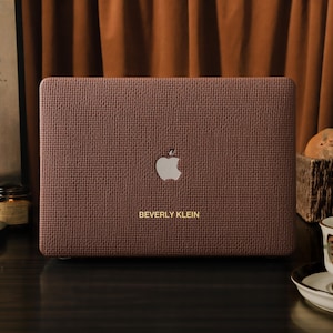 May include: A brown laptop case with a textured finish, displaying the Apple logo and the name "BEVERLY KLEIN" in gold. The case is set against a warm brown background, creating a stylish and elegant look.