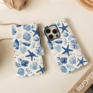 Puede incluir: Dos fundas blancas para teléfono con un estampado de conchas marinas y estrellas de mar azules. Una funda está abierta, revelando la cámara del teléfono. Las fundas están sobre una superficie clara.