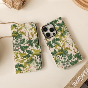 Może przedstawiać: Dwa etui na telefon z botanicznym wzorem. Jedno to etui w stylu folio z magnetycznym zamknięciem, a drugie to standardowe etui. Oba mają kremowe tło z zielonymi liśćmi i różowymi akcentami kwiatowymi. Etui są ozdobione kwiatowym wzorem.