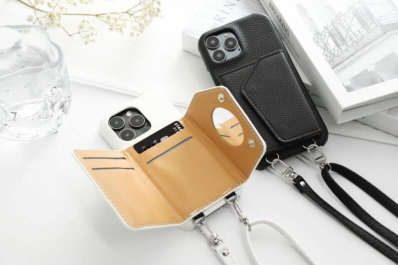 May include: Two white phone cases with a wallet and a strap. One case has a black leather exterior and the other has a tan leather interior. Both cases have a mirror on the back.
