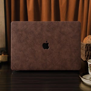 Könnte beinhalten: Eine braune Laptop-Hülle mit einem strukturierten, krokodilartigen Muster. Die Hülle hat eine Aussparung für das Apple-Logo. Der Hintergrund umfasst einen braunen Vorhang, ein Gemälde und eine Teetasse.