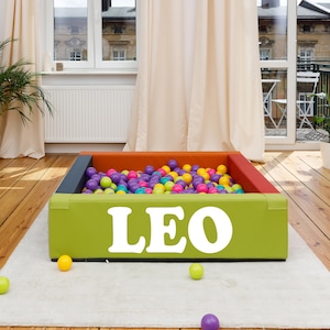 Könnte beinhalten: Ein rechteckiges Bällebad mit dem Namen "LEO" in weißen Buchstaben auf der Vorderseite. Das Becken ist mit bunten Bällen gefüllt und hat ein grünes, orangefarbenes, rotes und graues Farbschema. Das Bällebad steht auf einem weißen Teppich.