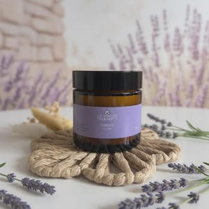 Peut inclure: Un petit pot en verre ambré avec un couvercle noir et une étiquette violette portant le texte LA BOUGIE THERHAPPY HARMONIE Lavande. Le pot repose sur un dessous de verre tressé beige, entouré de brins de lavande séchée sur une surface blanche.