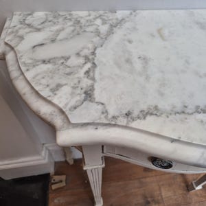 May include: A semi-circular console table featuring a white marble top with grey veining and a scalloped edge. The table has a light-coloured wooden base with fluted legs and a single drawer front adorned with a small, circular metal pull handle.