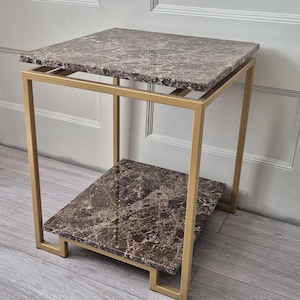 May include: A contemporary side table featuring a square top and a lower shelf with a marble-effect finish. The surfaces display a brown and beige veined pattern. The gold-coloured frame adds a touch of elegance. The table is approximately 50 cm wide and 50 cm deep.