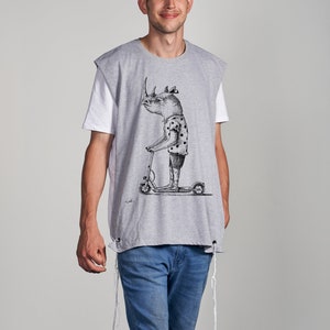 Peut inclure: Un t-shirt gris sans manches avec une illustration en noir et blanc d'un rhinocéros conduisant une trottinette. Le t-shirt a des cordons blancs qui pendent du bas.