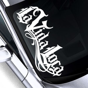 Può includere: Adesivo in vinile bianco con le parole "La Vandalora" in un font gotico, applicato al finestrino di un'auto. L'adesivo ha un design decorativo e fluido.