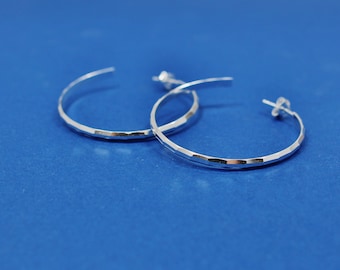 May include: A pair of silver hoop earrings with a textured, hammered finish. The earrings are on a blue background.