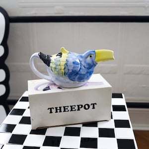 Könnte beinhalten: Eine skurrile Keramik-Teekanne in Form eines blauen Vogels mit gelbem Schnabel, platziert auf einer Box mit der Aufschrift "THEEPOT". Die Teekanne hat einen weißen Griff und ist mit blauen, gelben und schwarzen Details bemalt. Sie steht auf einer schwarz-weiß karierten Oberfläche.