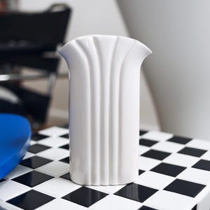 Könnte beinhalten: Eine weiße Keramikvase mit geriffeltem Design steht auf einer schwarz-weißen Schachbrettfläche.